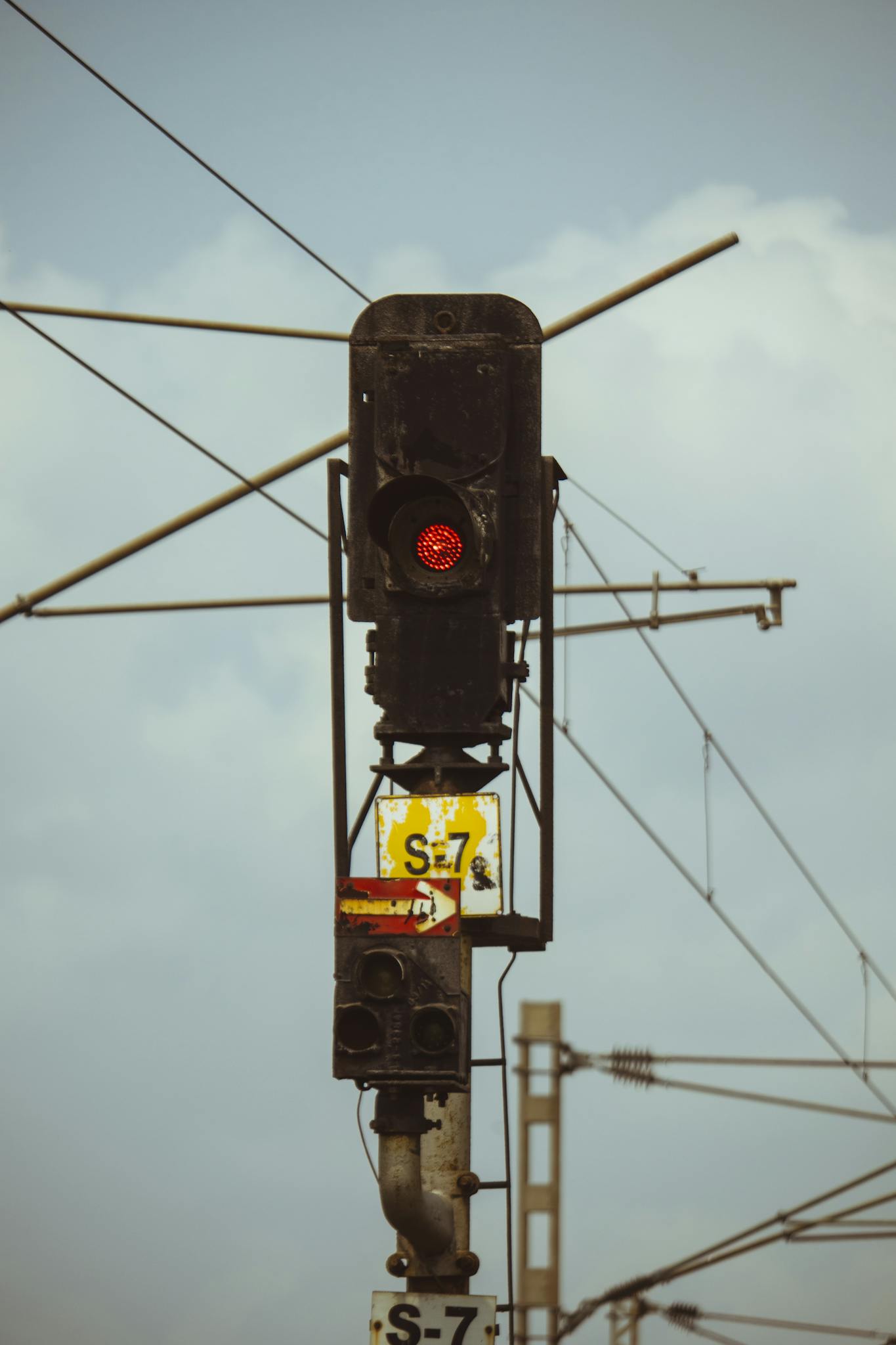 Close-up of a red railway signal with electrical wires and a cloudy sky in the background.
